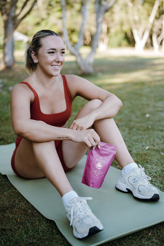 Woman sitting on an exercise mat in a park holding a pouch of Ritua Creatine for Women