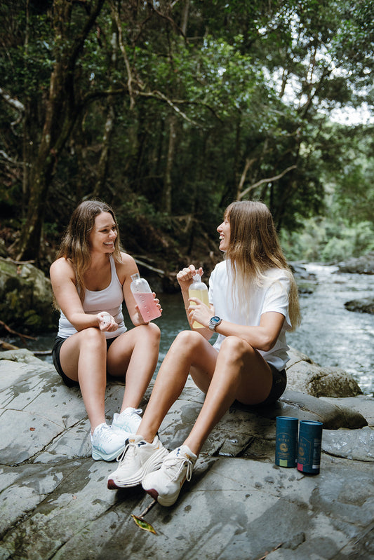 Two women sitting on a rock by a rainforest creek, sharing Ritua Hydrate in their drink bottles