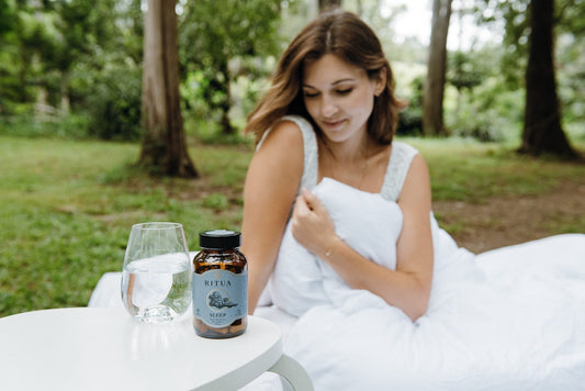 Woman wrapped in a white doona outdoors beside a table with a glass of water and a bottle of Ritua Sleep